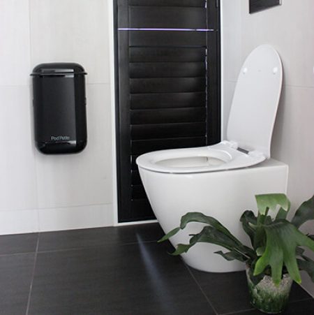 A black Pod Petite sanitary disposal unit beside toilet with a Pod Wrap Carbon Fibre decal.